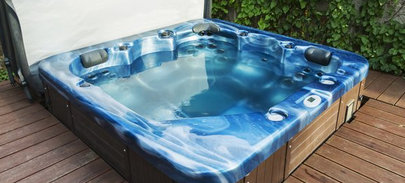 A blue, water-filled hot tub with built-in headrests is set on a wooden deck surrounded by greenery.